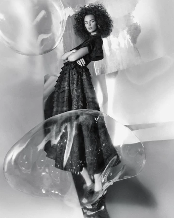Monochrome studio portrait of a woman with curly hair wearing a dark dress, standing among clear decorative bubbles and abstract shapes.