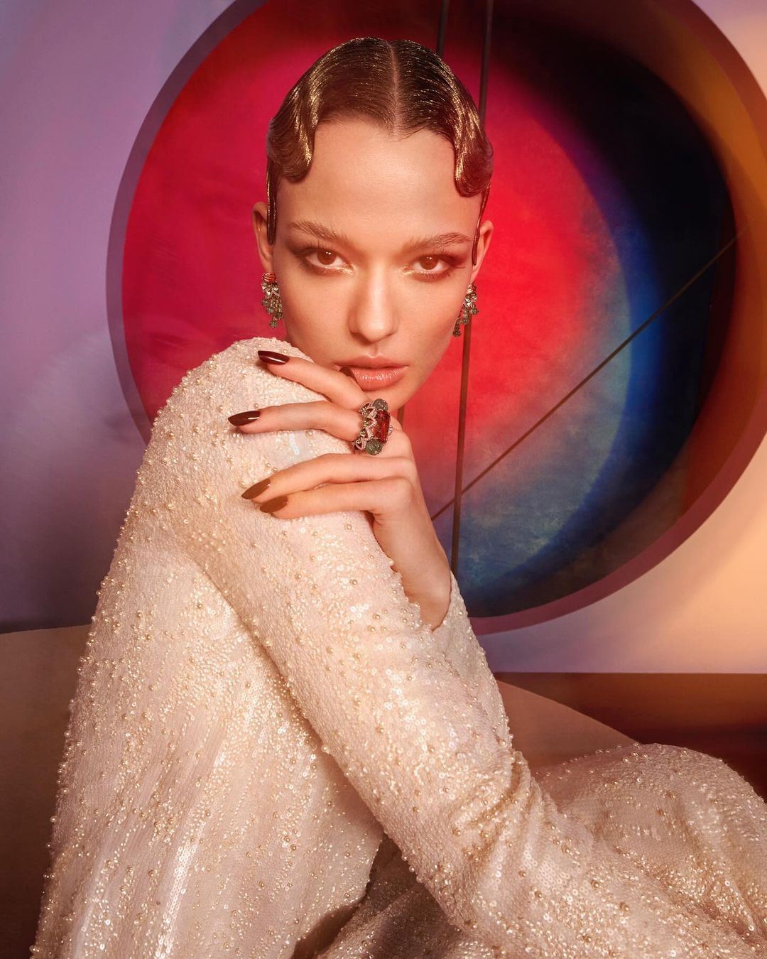 Close-up portrait of a young adult female model with wavy hairstyle, wearing an embellished gown, posing against a multicolored backdrop.