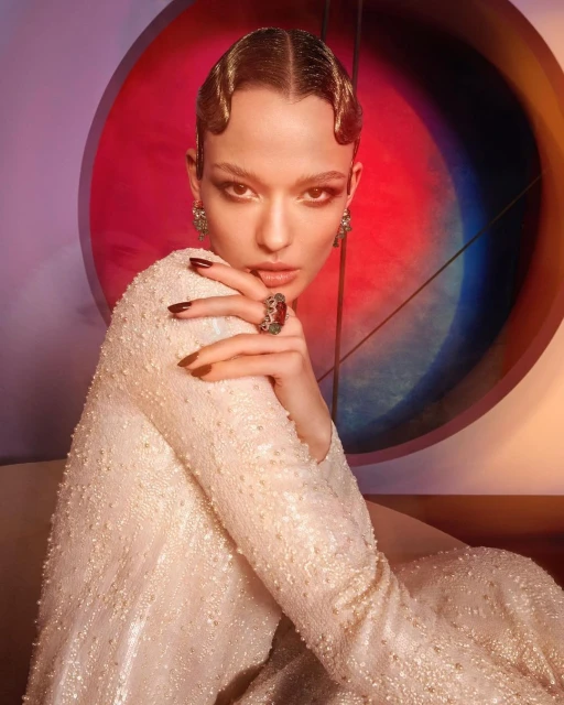 Close-up portrait of a young adult female model with wavy hairstyle, wearing an embellished gown, posing against a multicolored backdrop.