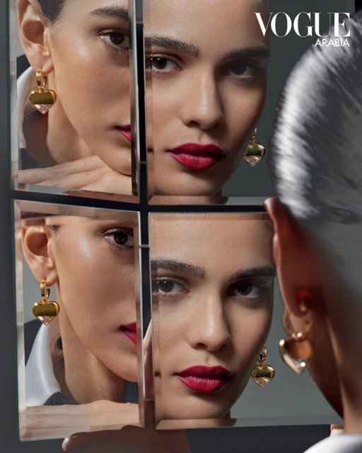 Portrait of a woman with dark hair reflected in a grid of mirrors wearing gold heart earrings and red lipstick, Vogue Arabia logo visible.