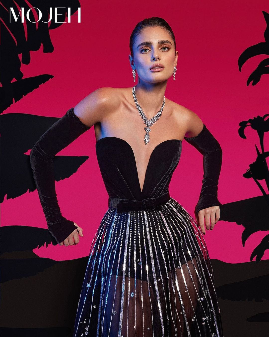 Taylor Hill wearing a black strapless gown with silver embellishments and long black gloves against a magenta backdrop