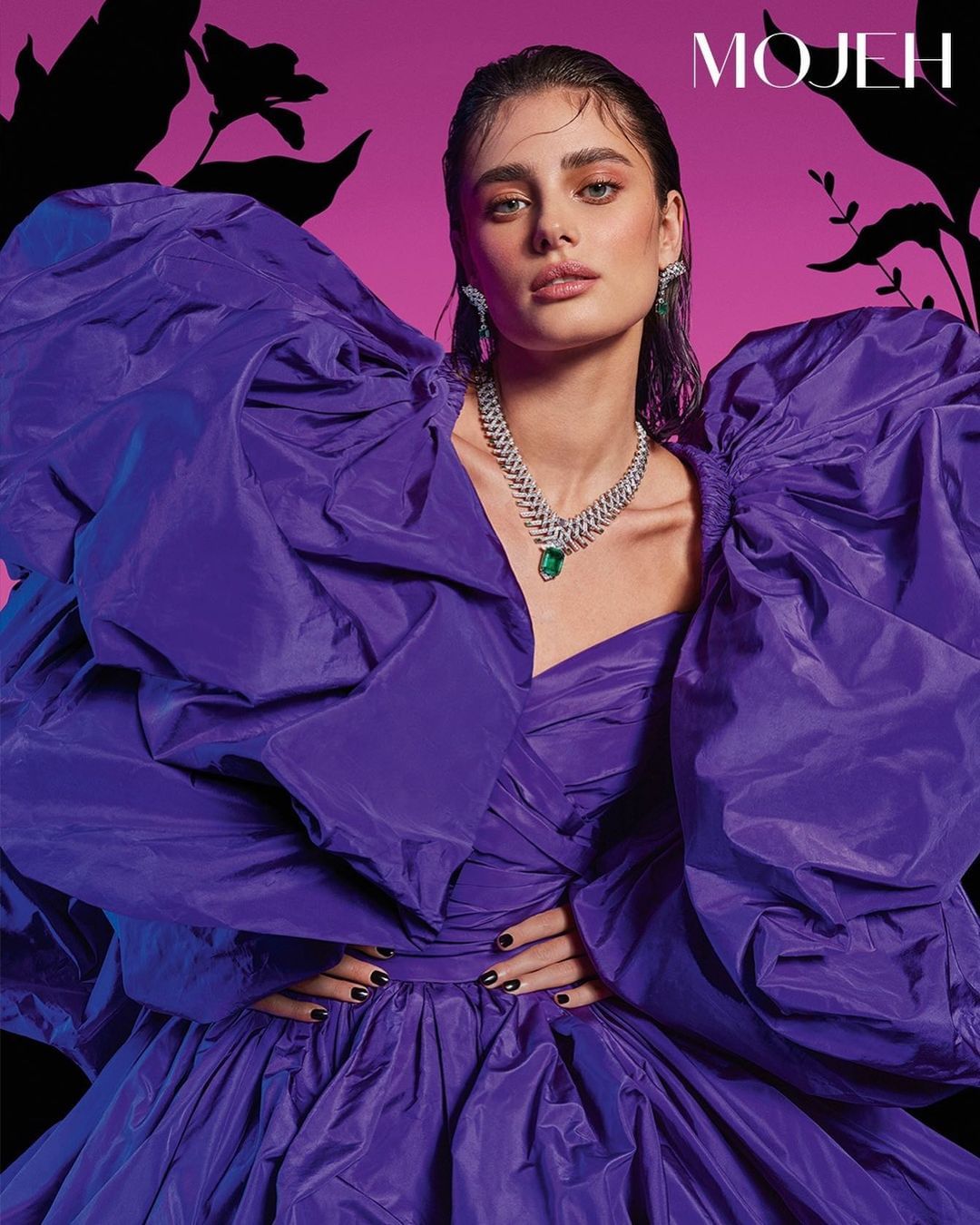 Fashion portrait of a woman wearing a large purple dress and jewelry posing against a pink and black floral background.