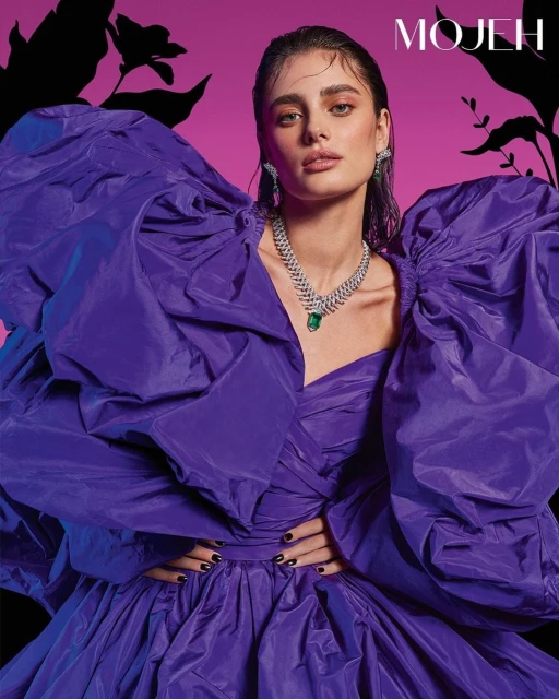 Fashion portrait of a woman wearing a large purple dress and jewelry posing against a pink and black floral background.
