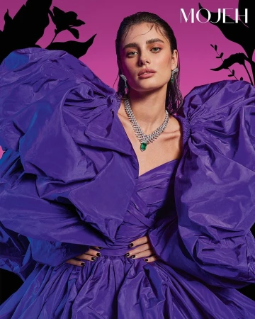 Fashion portrait of a woman wearing a large purple dress and jewelry posing against a pink and black floral background.