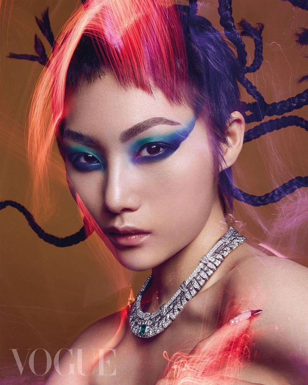 Close-up studio portrait of a striking woman with red and purple hair, bold makeup, and an elegant diamond necklace against a golden background.