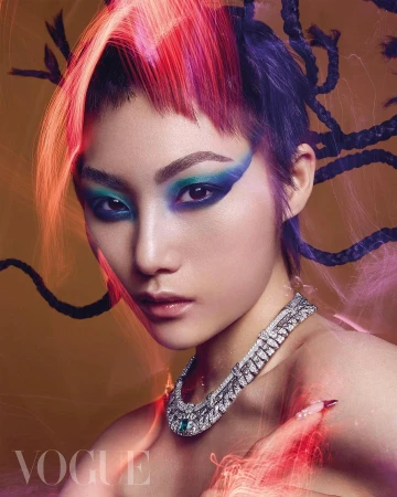 Close-up studio portrait of a striking woman with red and purple hair, bold makeup, and an elegant diamond necklace against a golden background.