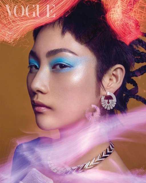 Close-up of an Asian woman wearing blue eyeshadow, a patterned necklace, and earrings in a Vogue cover shoot with orange strands.
