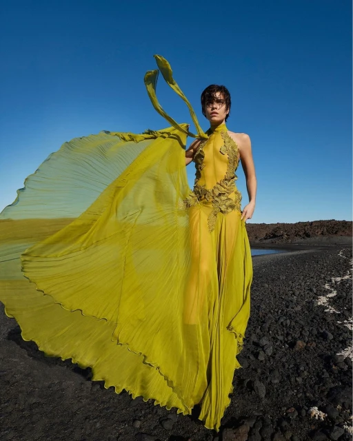 Woman in flowing yellow dress poses on black rocky landscape under a bright blue sky. Fashion editorial with natural light.