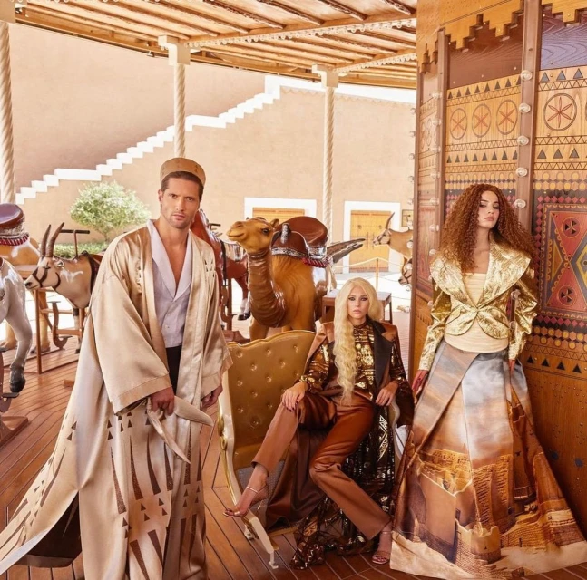 Three people pose wearing Arabian-inspired clothing, with animal carousel figures of a camel, oryx, and horse in a tan-colored interior.