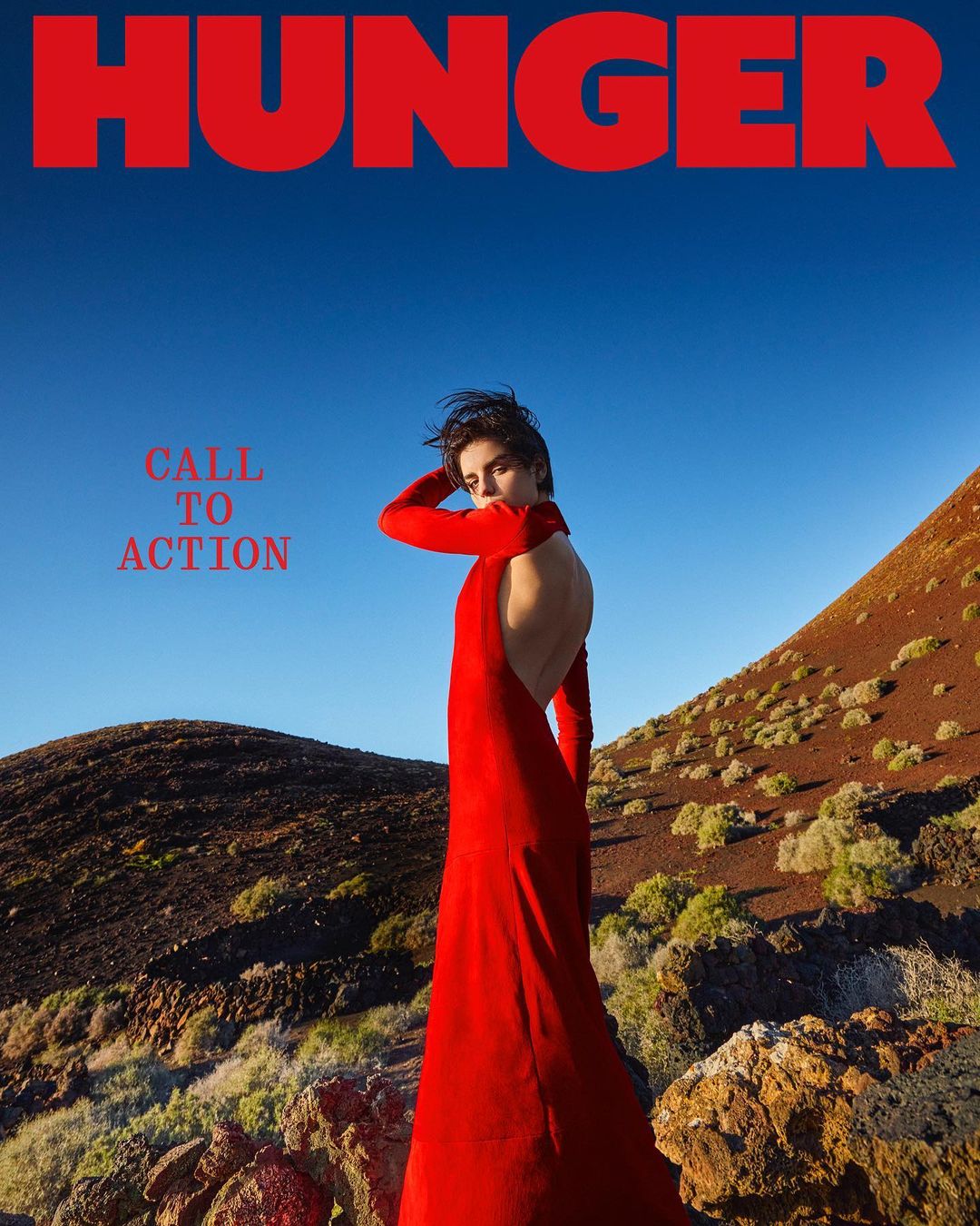 Model in a long red dress poses on rocky terrain under a blue sky, promoting Hunger magazine with a 'Call to Action' message.