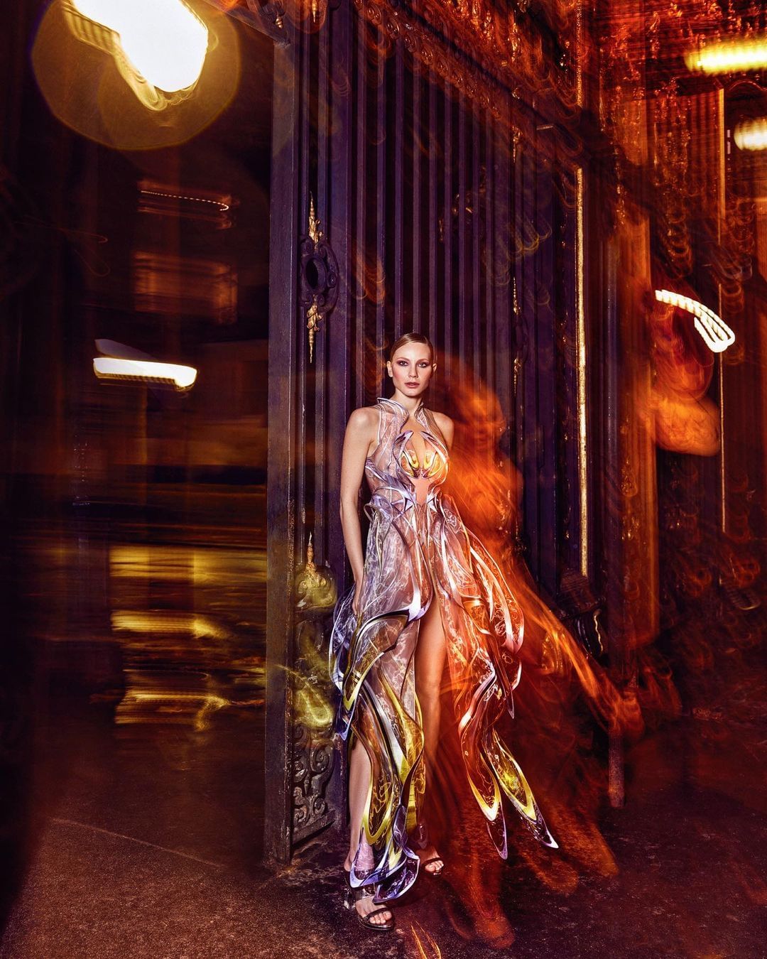 Elegant adult female poses in a flowing gown near an ornate metal gate with diffused golden lighting and a long exposure effect.