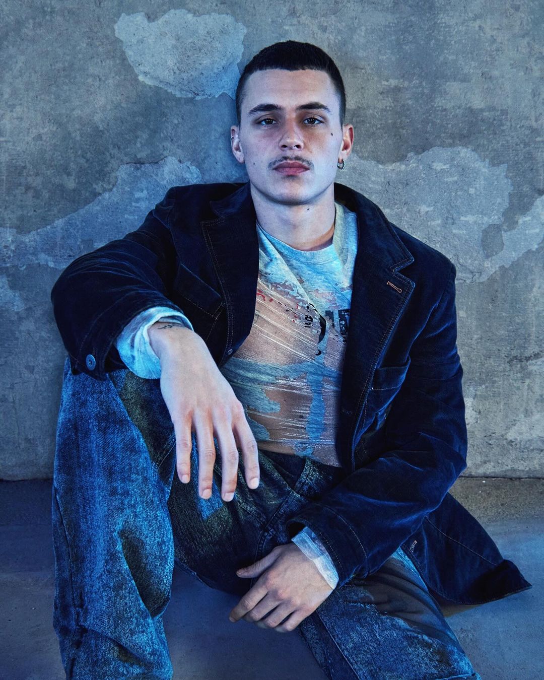 Portrait of a young adult male in a casual pose against a textured concrete wall, wearing a velvet blazer and patterned jeans.
