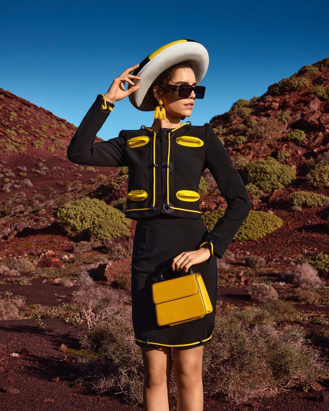 A fashionable woman in a black skirt suit with yellow trim and a wide-brimmed hat poses in a desert landscape under a clear blue sky.