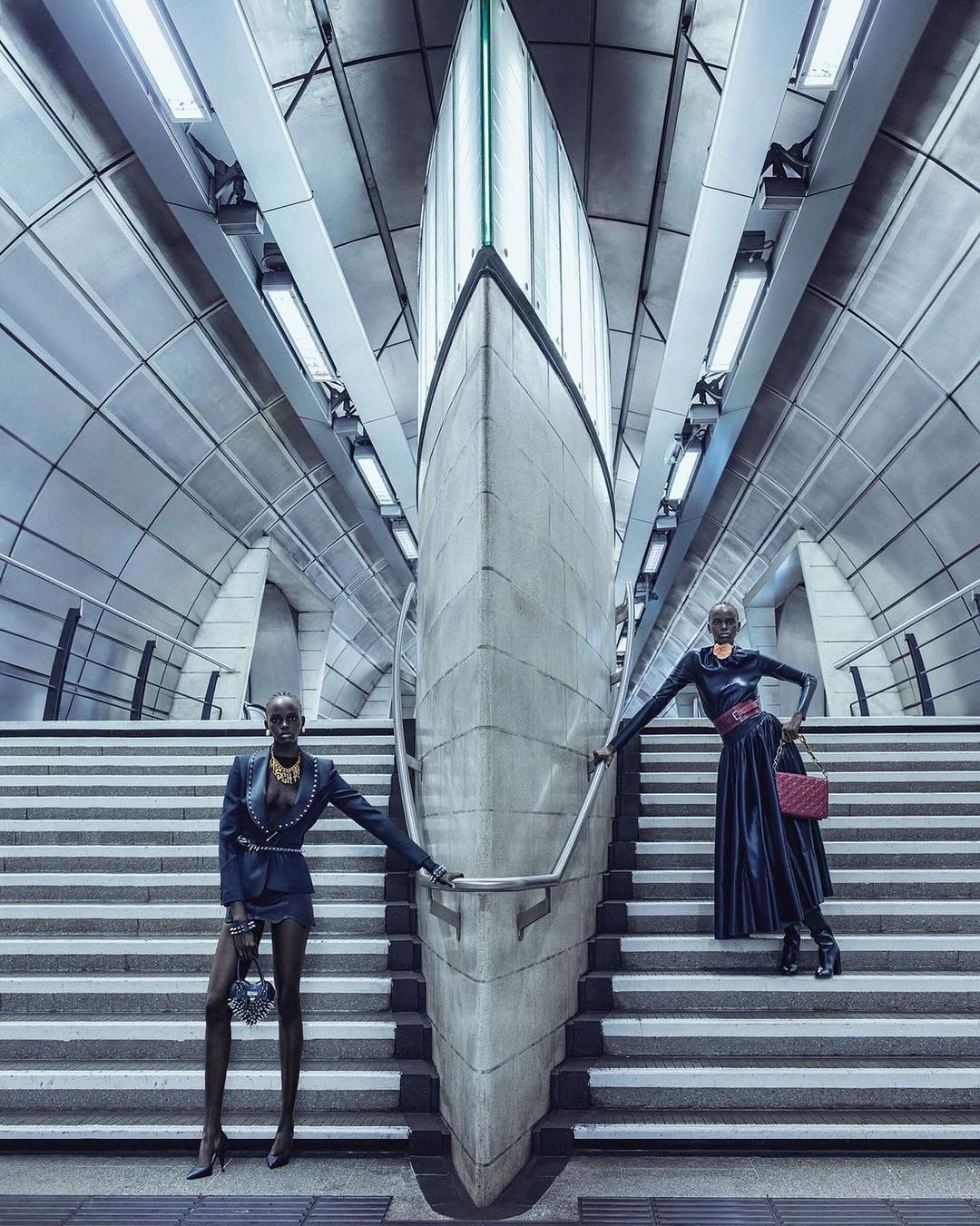 Two women in stylish black outfits pose on gray steps of an underground station, symmetrically framed by modern architecture.