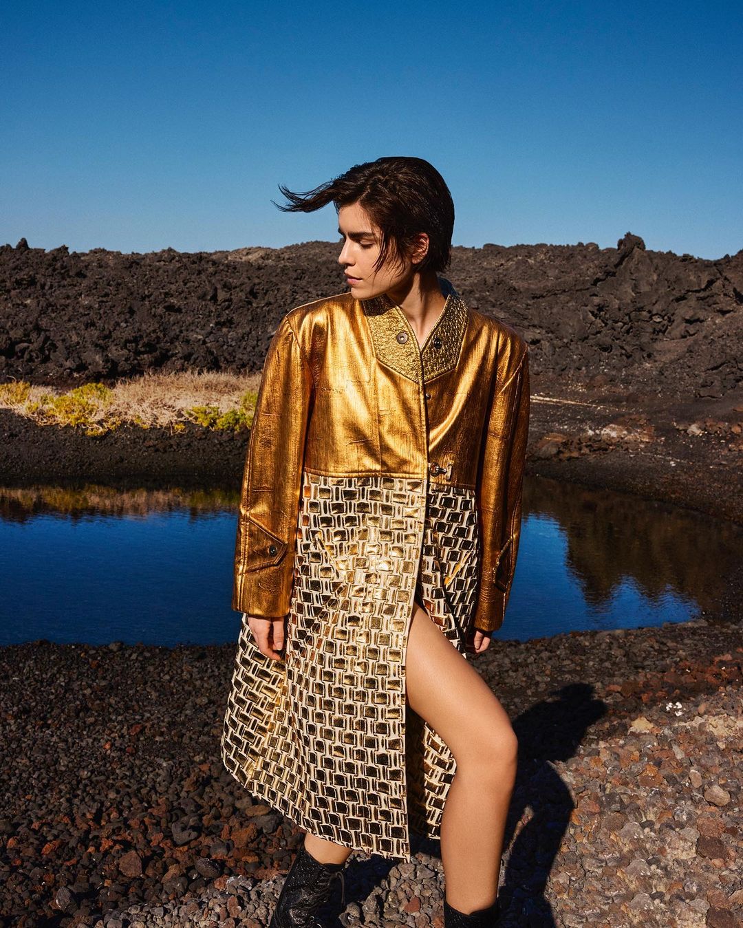 A woman with short brown hair poses outdoors in a shiny gold and patterned coat against a rocky landscape on a sunny day.