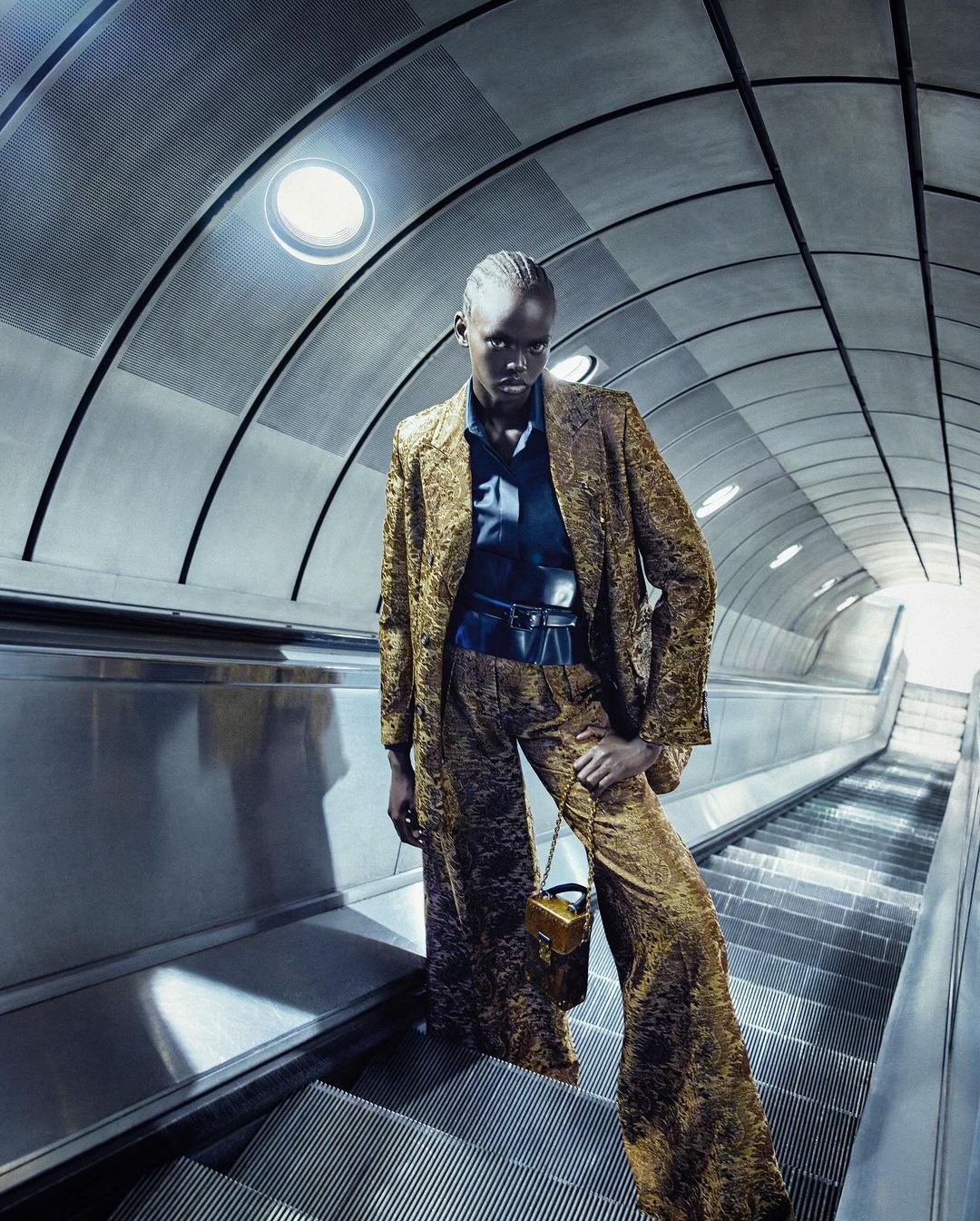 Fashionable black woman poses in gold patterned suit with a purse on an escalator in an underground subway station, editorial shot.