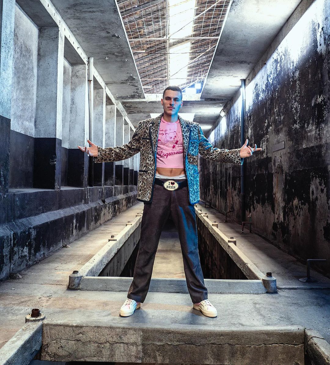 A young adult male in a leopard print jacket and pink shirt strikes a pose in a concrete structure with linear design elements.