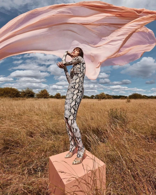 Adult female fashion model in a snake-print dress on a cube in a field with a flowing pink scarf against a blue sky backdrop.