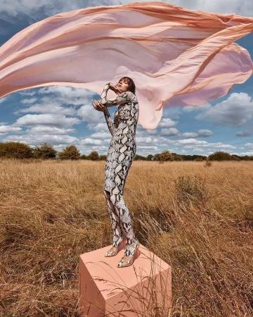 Adult female fashion model in a snake-print dress on a cube in a field with a flowing pink scarf against a blue sky backdrop.