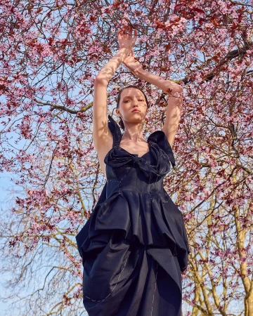 Woman in a dark dress with arms raised stands under pink blossoms of a flowering tree against a blue sky on a sunny day.