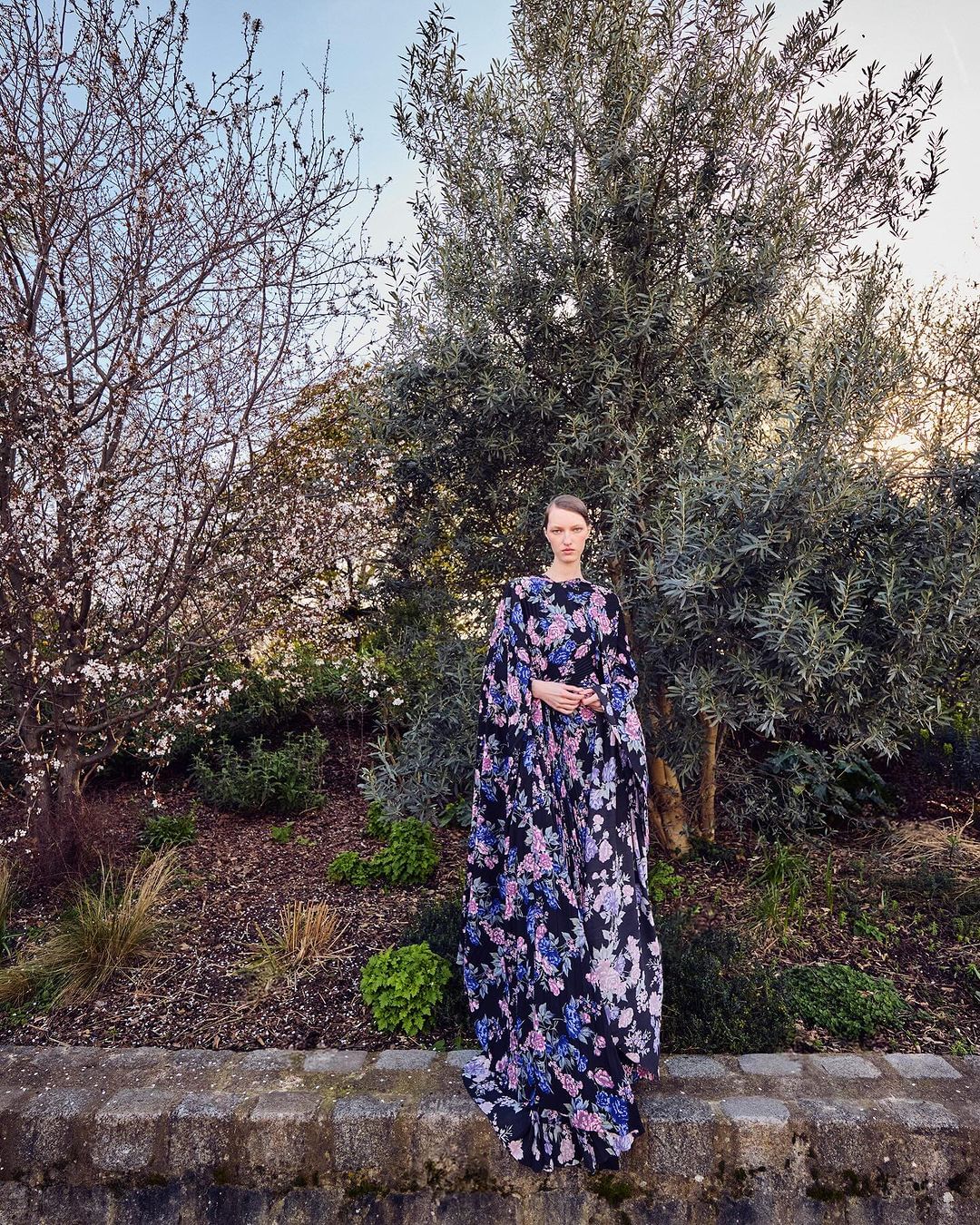 A stylish woman stands outdoors wearing a long floral dress with a garden and trees in the background on a sunny day.