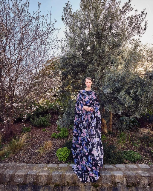 A stylish woman stands outdoors wearing a long floral dress with a garden and trees in the background on a sunny day.