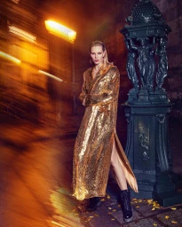 A blond woman wearing a long gold sequin coat posing next to a dark ornate streetlight in Milan at night.