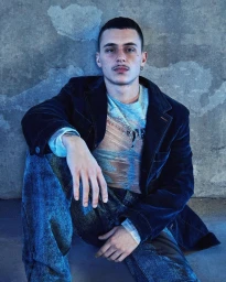 Portrait of a man in a dark blue corduroy jacket and patterned t-shirt seated against a textured concrete wall.