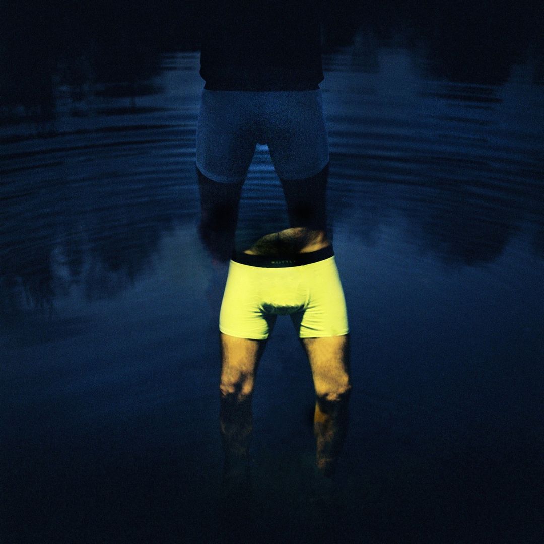Man stands waist-deep in dark water, wearing white boxers with reflection showing yellow boxers, creating a surreal, mirrored effect in nature.