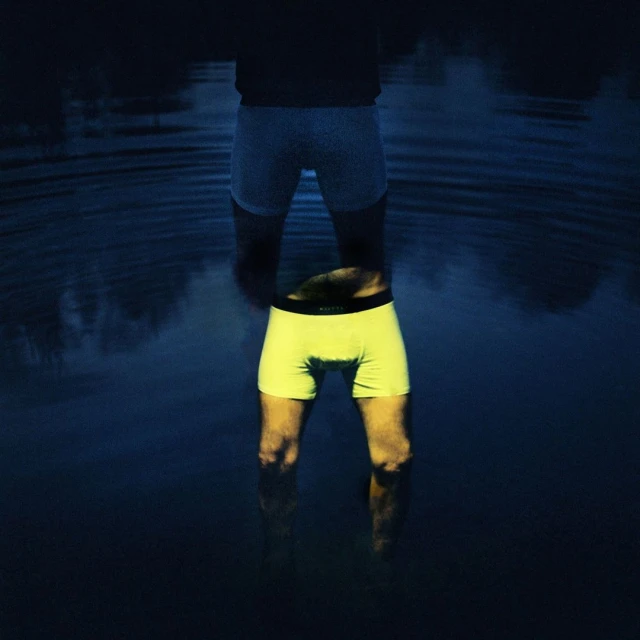 Man stands waist-deep in dark water, wearing white boxers with reflection showing yellow boxers, creating a surreal, mirrored effect in nature.