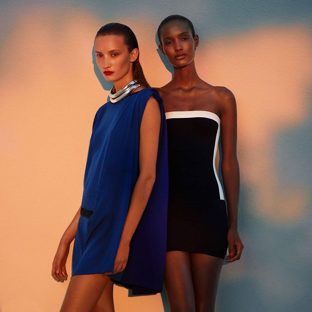Two women in studio fashion shot. One woman wears a blue dress and silver collar necklace. The other wears a black and white dress.