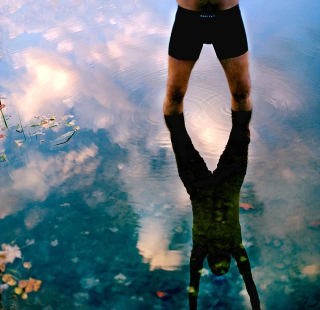 Torso and legs of an adult male standing in water with sky reflections. The man has a darker reflection in the water.
