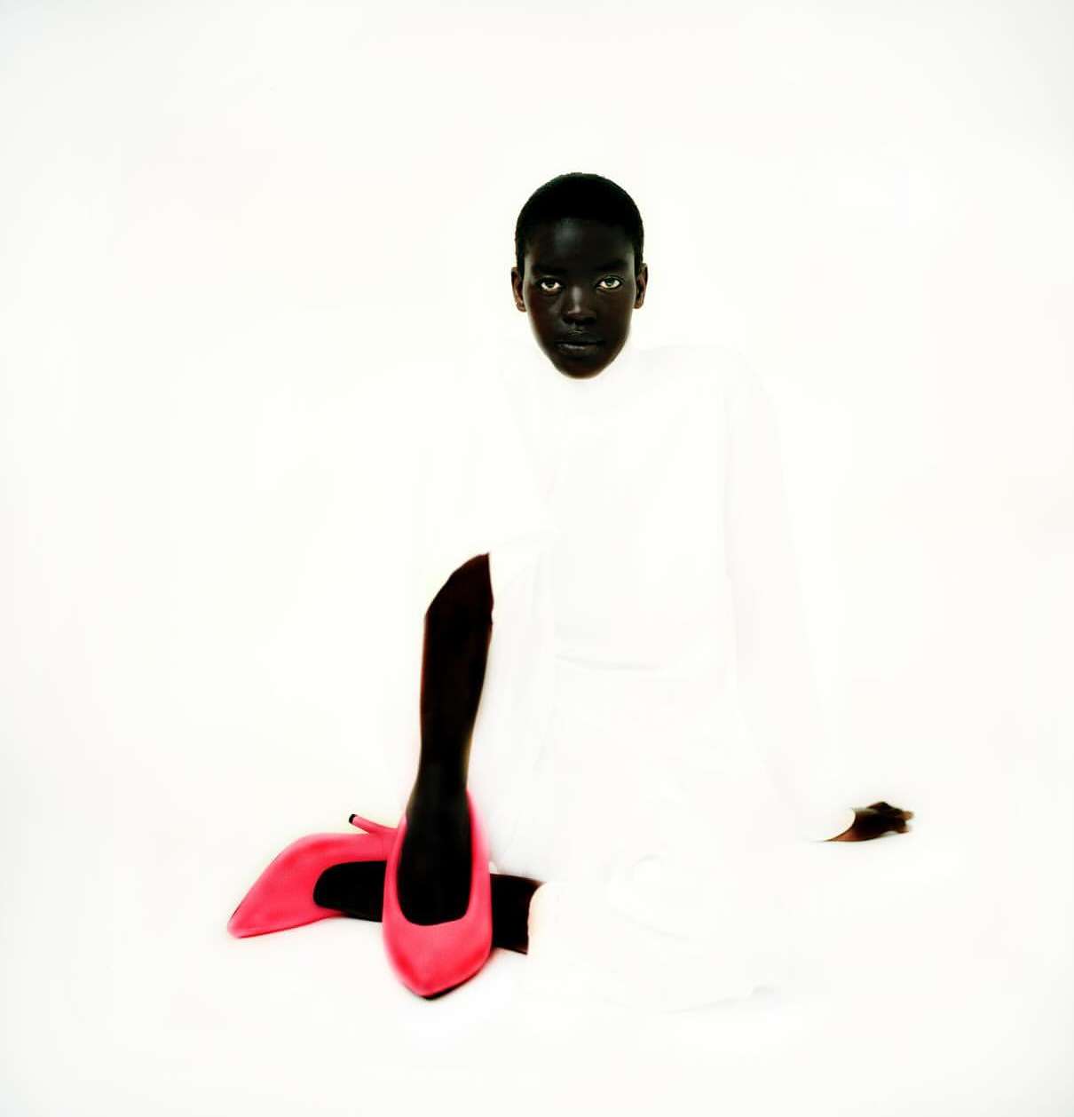 A dark-skinned person in a white garment sits on a white backdrop with a pair of pink high heel shoes in the foreground.