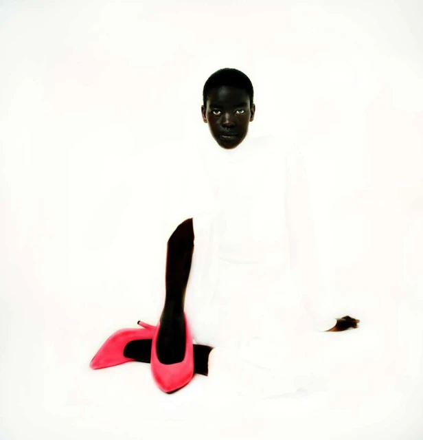 A dark-skinned person in a white garment sits on a white backdrop with a pair of pink high heel shoes in the foreground.