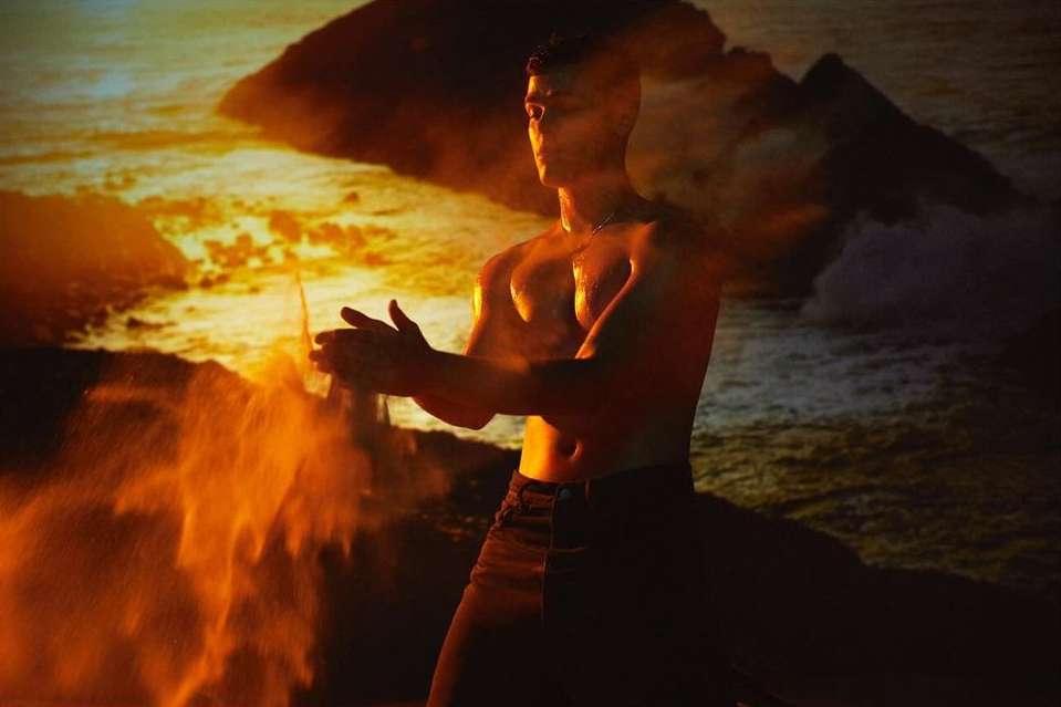 Man with a bare chest throwing sand at a golden beach with a soft dreamy feel. He is dark-haired and wears dark pants.