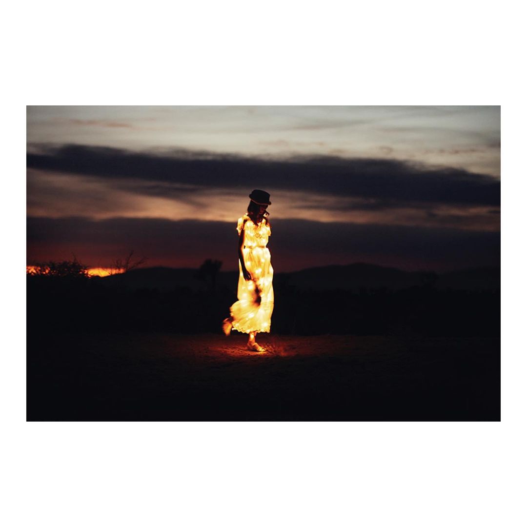 Woman in a long dress and a bowler hat illuminated by warm light poses in a field with a dark and cloudy sunset sky in the background.