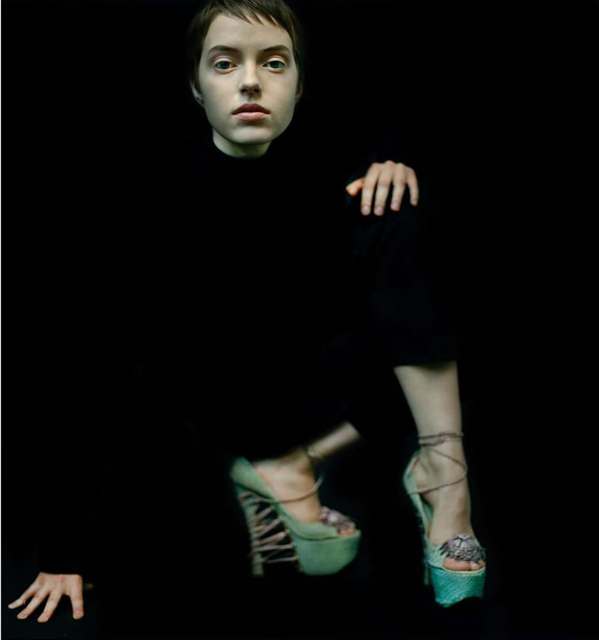 Fashion shot of a young woman with short hair wearing high-heeled platform shoes against a black background, focusing on pose and avant-garde style.