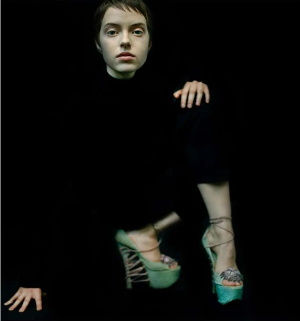 Fashion shot of a young woman with short hair wearing high-heeled platform shoes against a black background, focusing on pose and avant-garde style.
