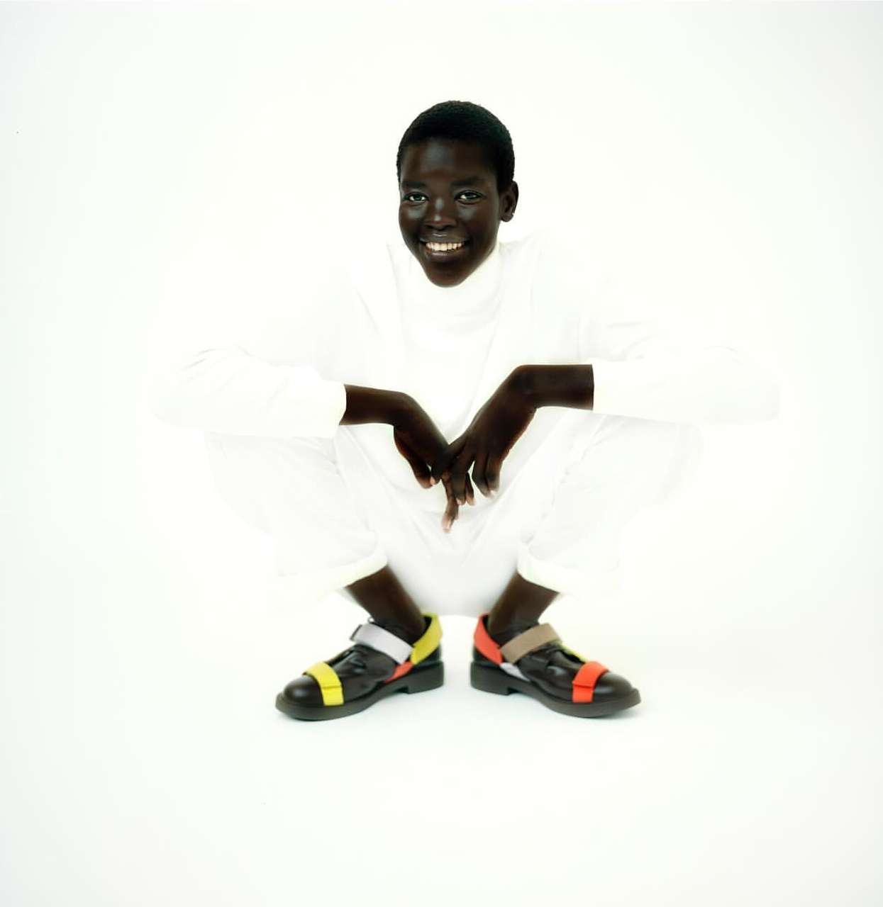 A smiling Black female adult sits cross-legged on a white background wearing a white outfit and black strappy shoes with yellow and orange accents.