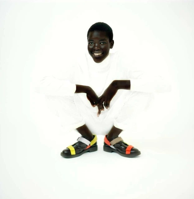 A smiling Black female adult sits cross-legged on a white background wearing a white outfit and black strappy shoes with yellow and orange accents.