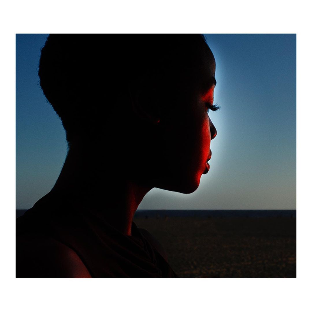 Profile of an adult female with short hair, silhouetted against a blue sky and horizon, with red light highlighting facial features.
