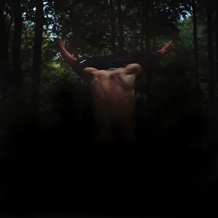 A shirtless adult male stands with his back to the viewer, partially undressed, with arms raised in a forest setting.