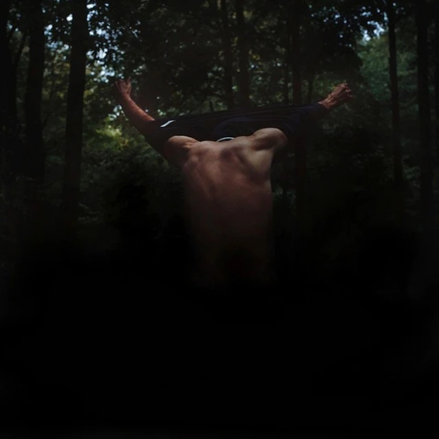 A shirtless adult male stands with his back to the viewer, partially undressed, with arms raised in a forest setting.