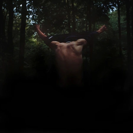 A shirtless adult male stands with his back to the viewer, partially undressed, with arms raised in a forest setting.