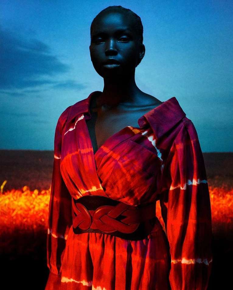 Black model poses in red and white tie-dye dress against a dusk background. The belt is red leather. The sky is a twilight blue.