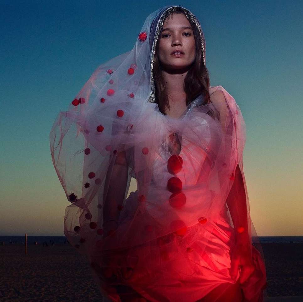 Fashion shot of a woman in a white dress with red dots, posing on a beach at dusk with a blue sky in the background, soft lighting.