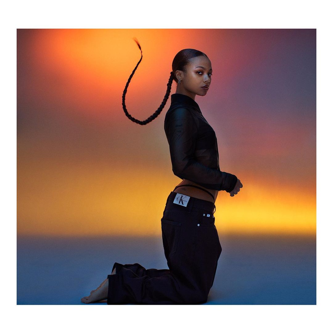 A Black female model with a long braid poses kneeling in a black top and jeans against a warm gradient background in a fashion shot.