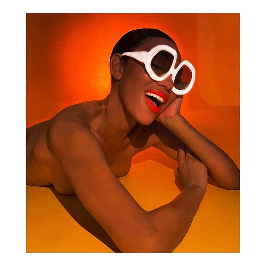 Smiling woman with short hair, bright red lipstick, and wearing oversized white sunglasses poses in front of an orange backdrop.