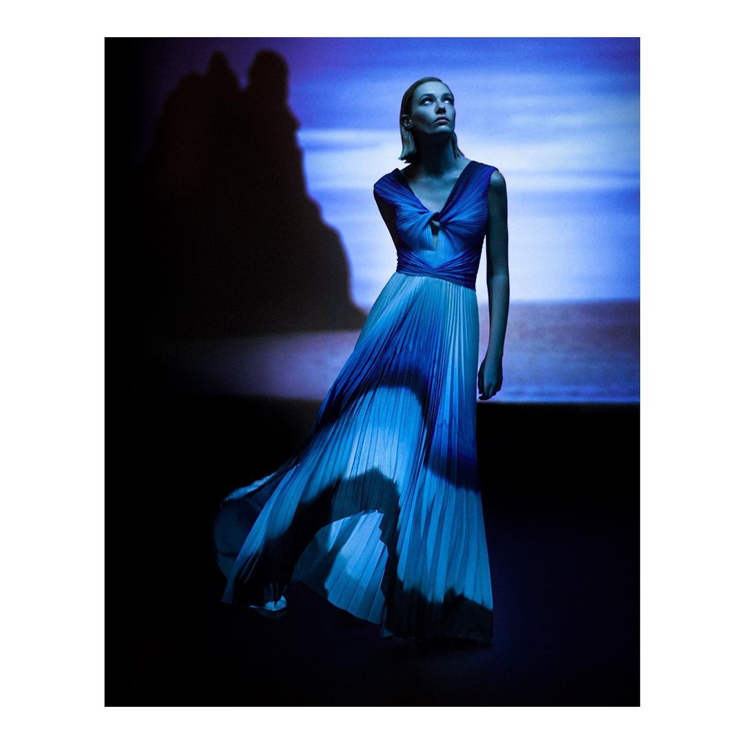 Fashion shot of a woman in a flowing ombre gown with a pleated skirt, posing against a background with an abstract landscape projection.