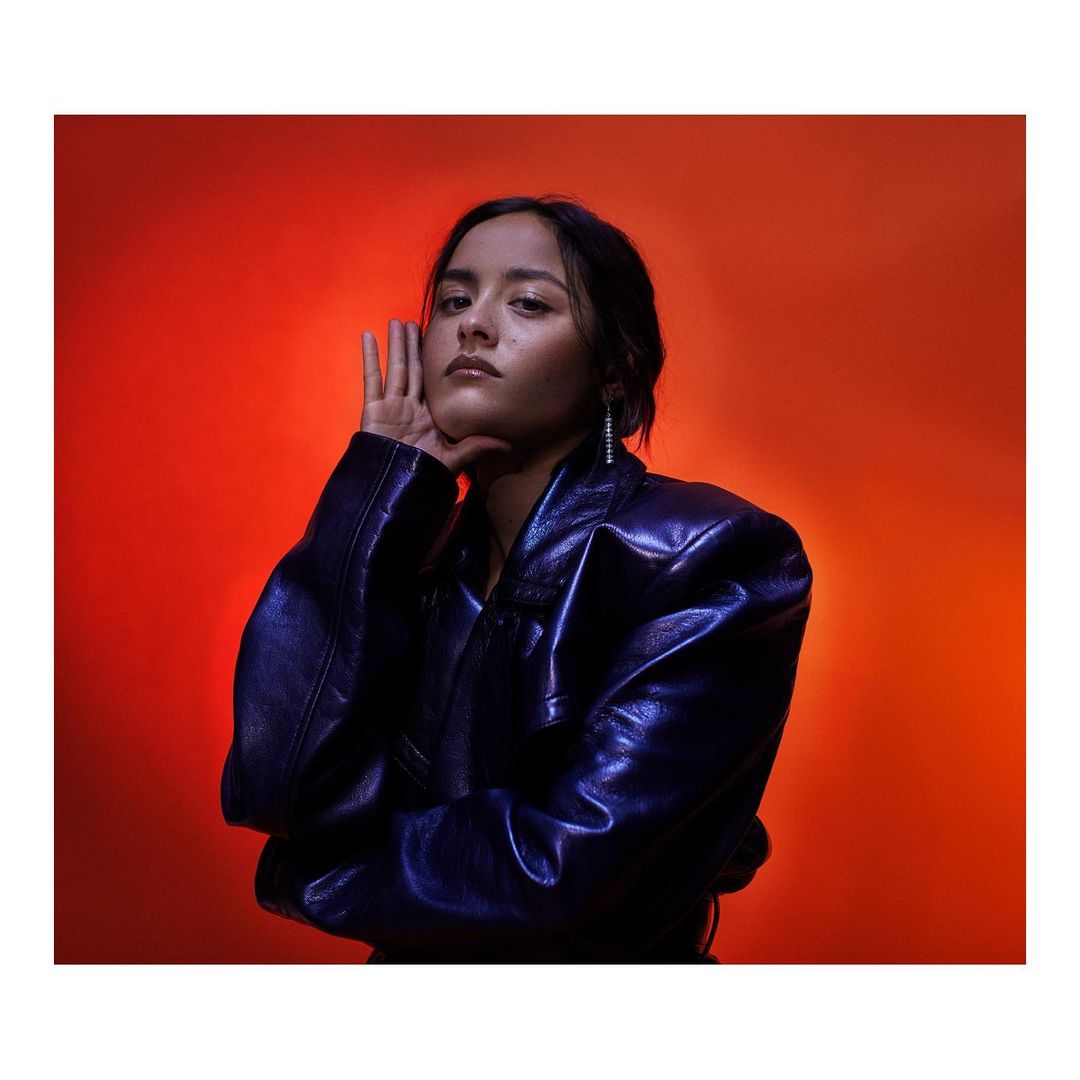 Portrait of a young adult female with a hand to her face, wearing a shiny dark-blue jacket against a warm gradient orange background.